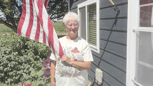 Byron Days Parade Grand Marshal Elaine Abraham stands on the front porch of her home, surrounded by colorful flowers and an American flag. Born in Canada, Abraham became a United States citizen in 2006.